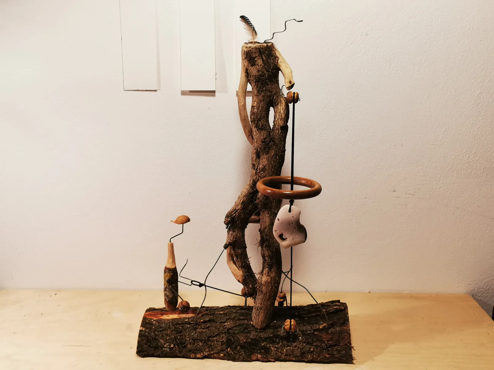 Scultura in legno con piuma, pietra forata e fili di ferro su base di scorza